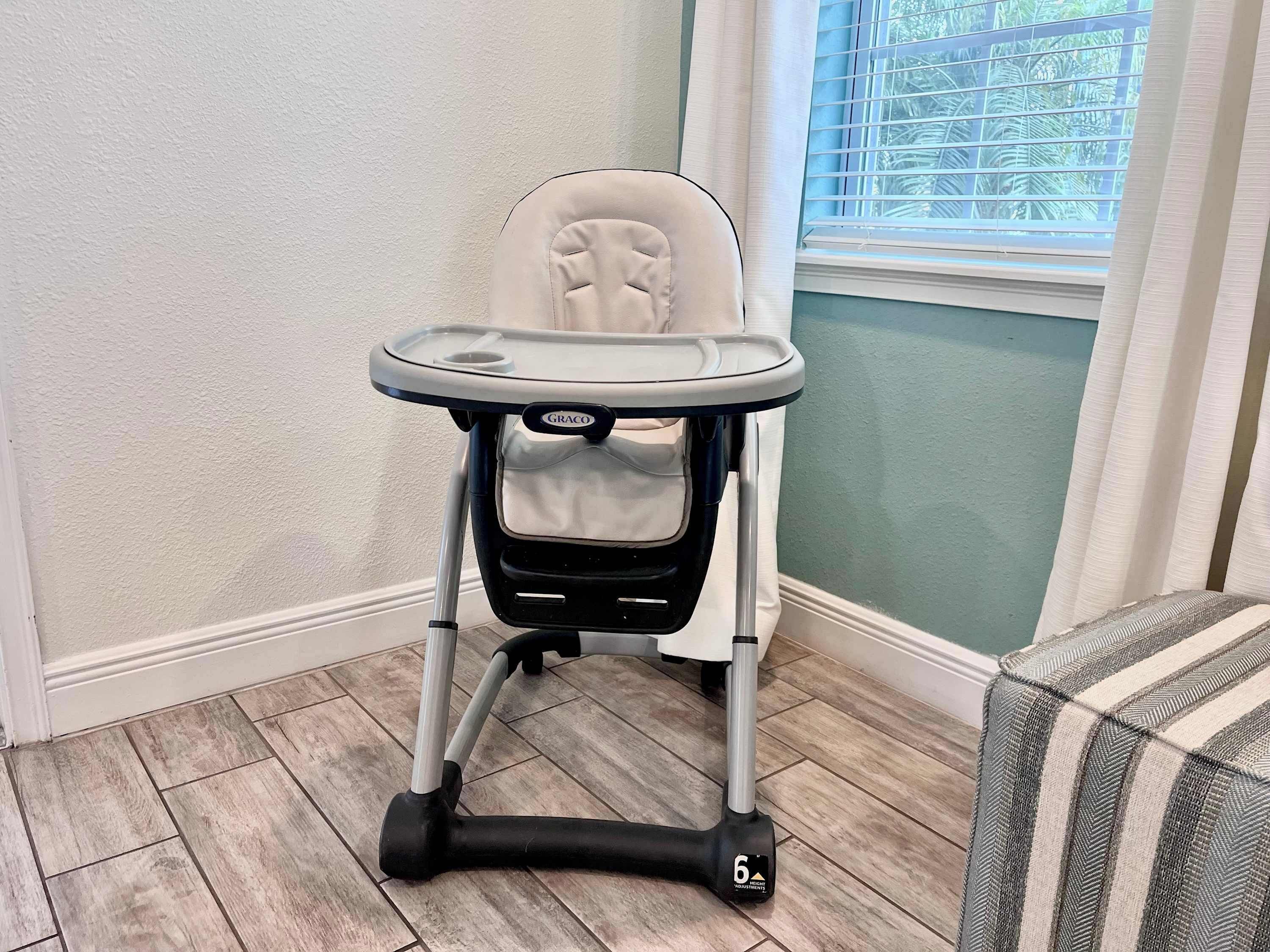A Graco high chair with a tray table, positioned in a corner of a room with wooden flooring and a window with blinds.