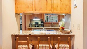 A kitchen with wooden cabinets, a granite countertop, a coffee maker, and a sink. There are three striped chairs at the counter. - Creekside - 2 BDR Resort Cabin in Epic Location! (Branson West)