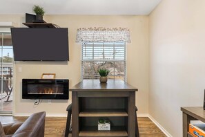 Un salon doté d’un téléviseur à écran plat fixé au mur, d’une cheminée, d’une fenêtre avec des stores et d’une table en bois sur laquelle est posée une plante en pot.