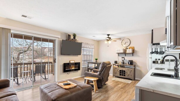 A modern living room with a sofa, ottoman, and a TV mounted on the wall. There is a dining area with a table and chairs, and a kitchen with a sink and cabinets. - Chandlers Pondview Getaway Chic Branson Escape (Branson)