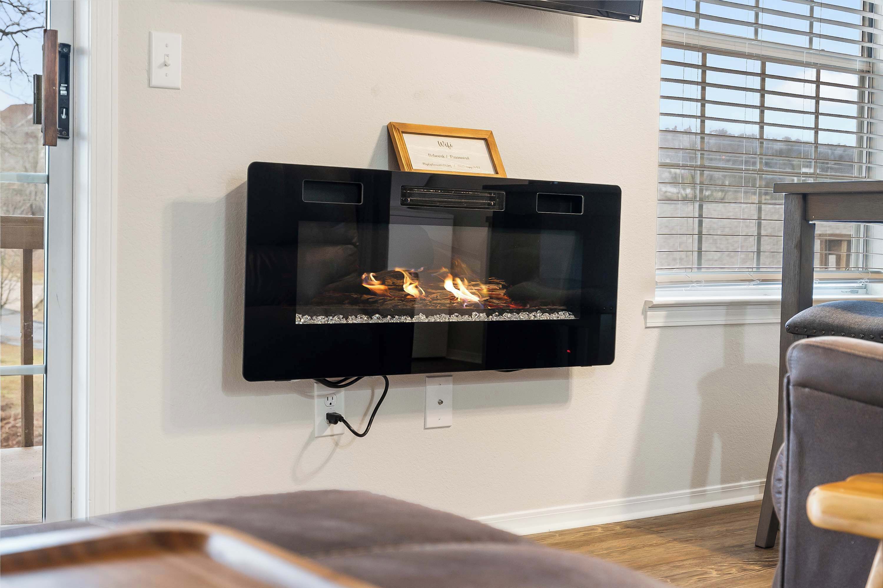 A wall-mounted fireplace with a framed certificate above it, a power outlet below, and a window with blinds to the right.