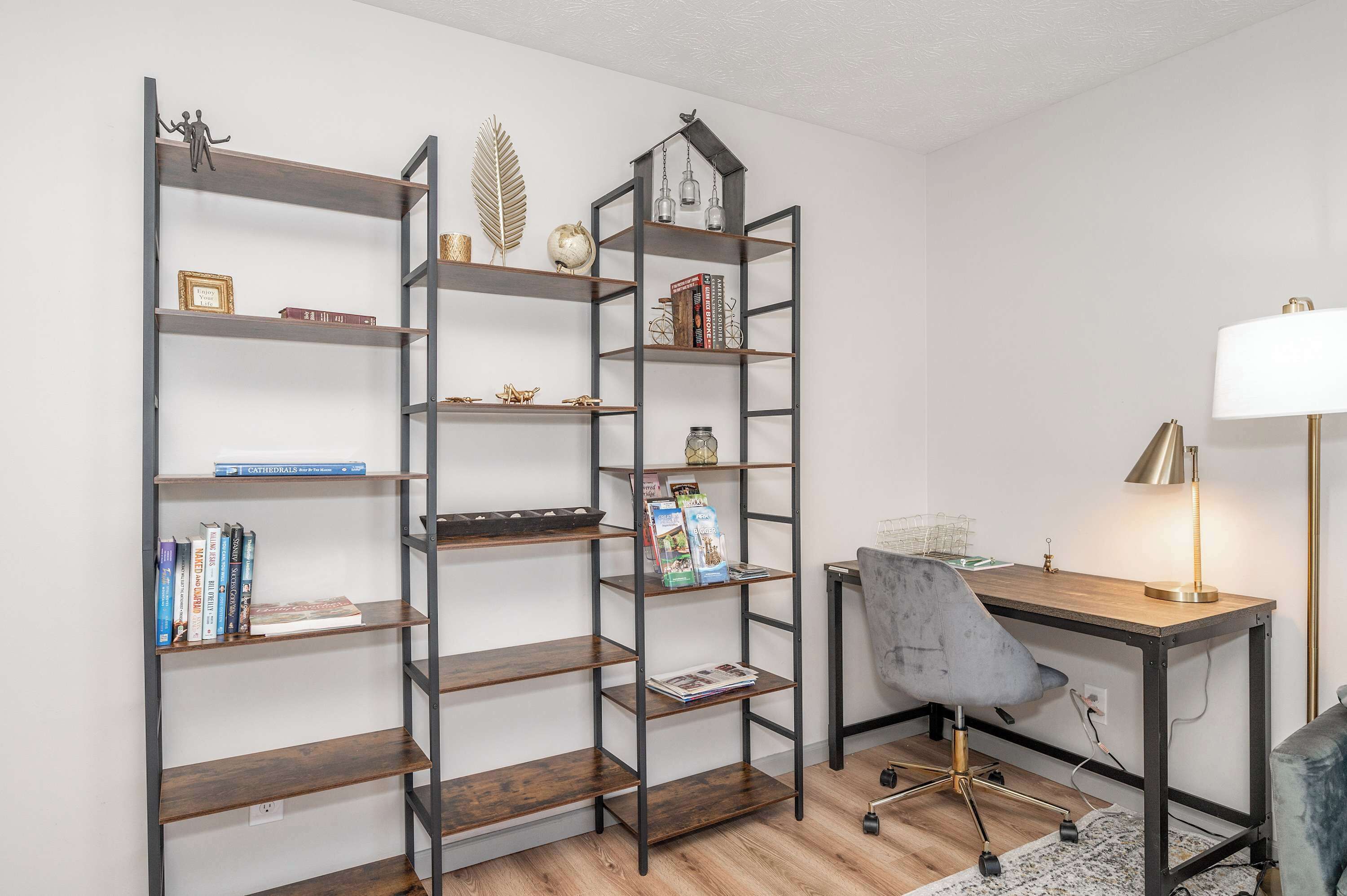 Ett rum med två höga, mörka träbokhyllor fyllda med böcker och dekorativa föremål, ett skrivbord med en lampa och en grå kontorsstol.