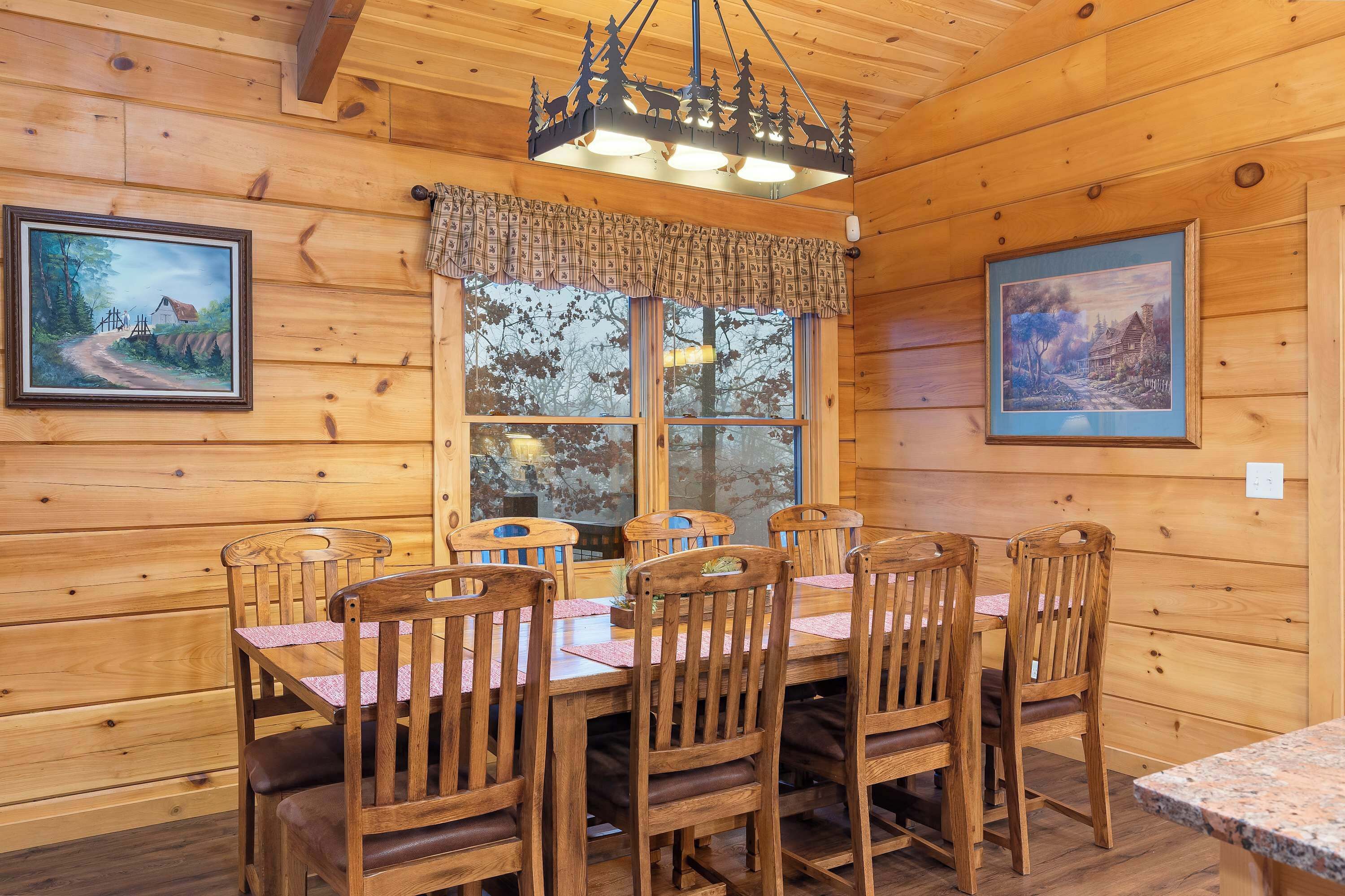 A wooden dining area with a table and chairs, a window with a view, and two framed paintings on the walls.