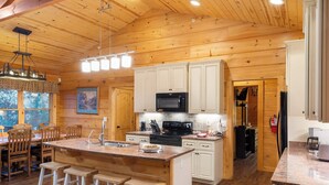 A wooden cabin interior with a kitchen featuring white cabinets, a central island with stools, a microwave, and a stove. - Teals Treasure Secluded Lakeside Cabin for 8! (Kirbyville)