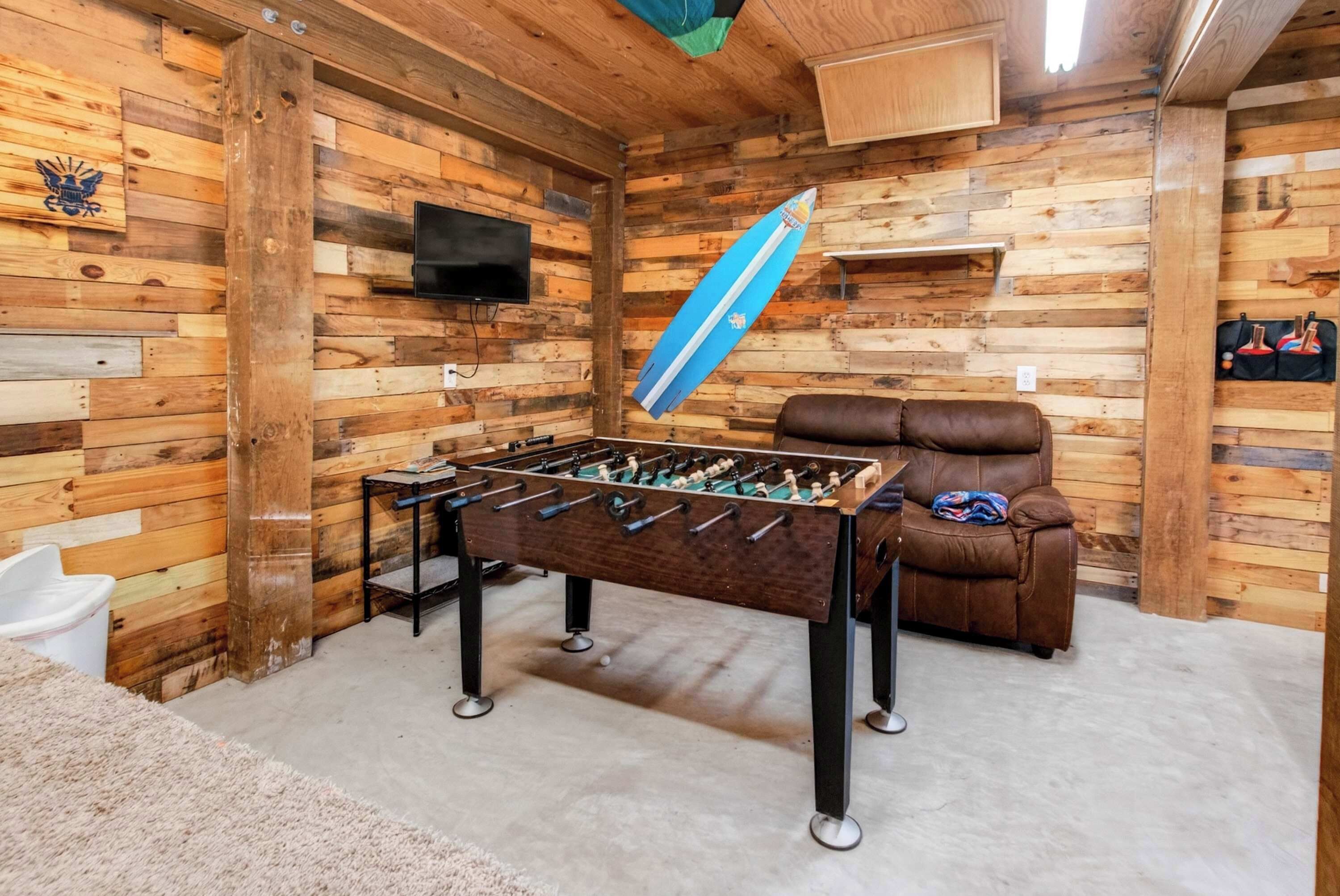 Una habitación con suelo de madera, una mesa de fútbol de mesa, un sofá de cuero marrón y un televisor montado.