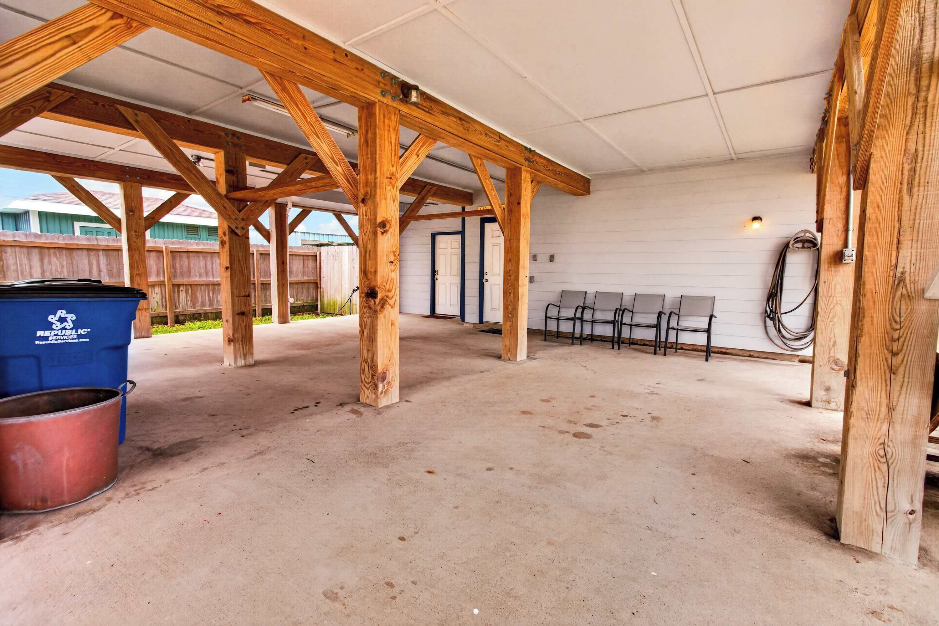 An open covered area with wooden beams, a concrete floor, and a blue trash bin.