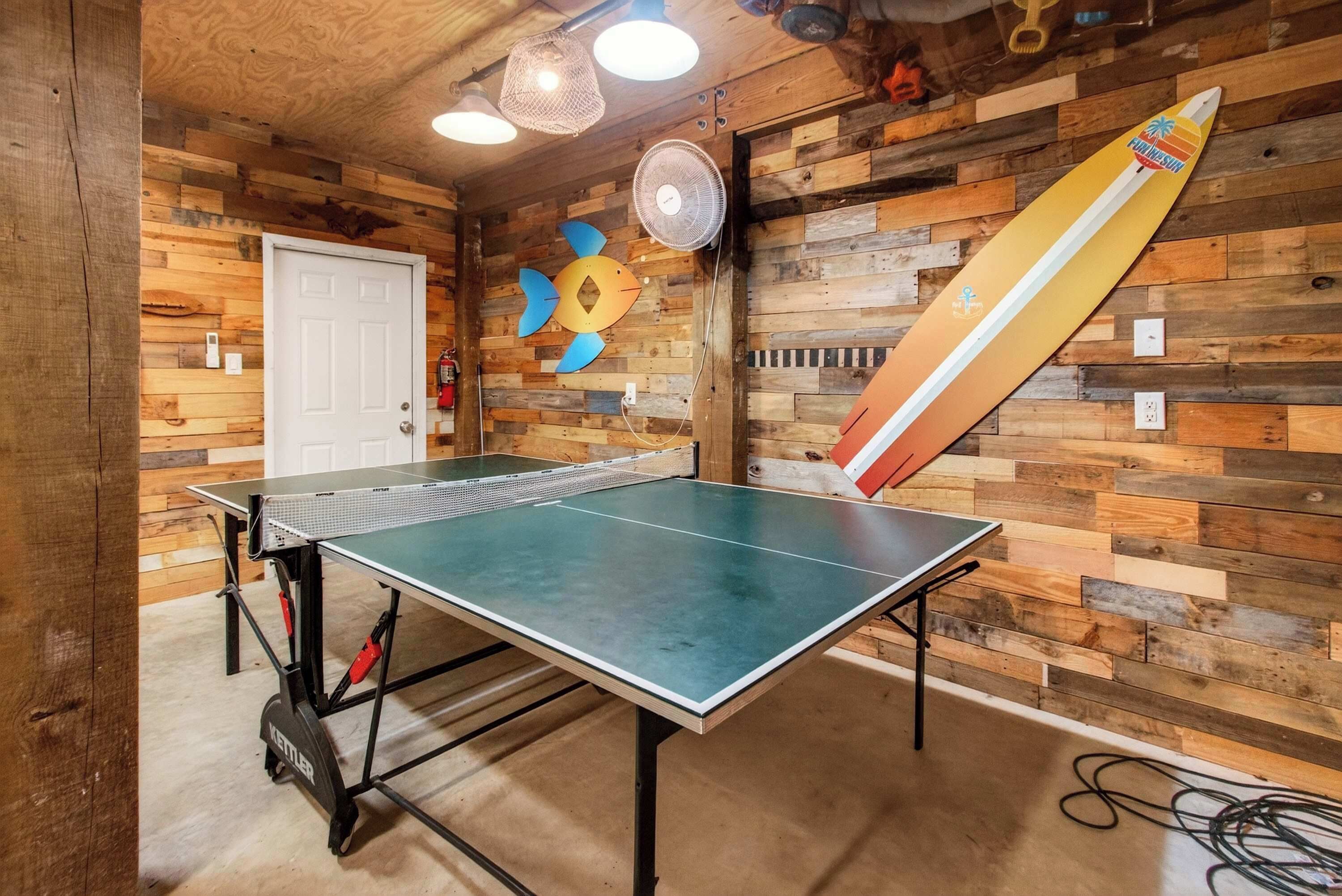 Una mesa de ping pong en una habitación con paredes de madera, un ventilador y una gran decoración con forma de lápiz.