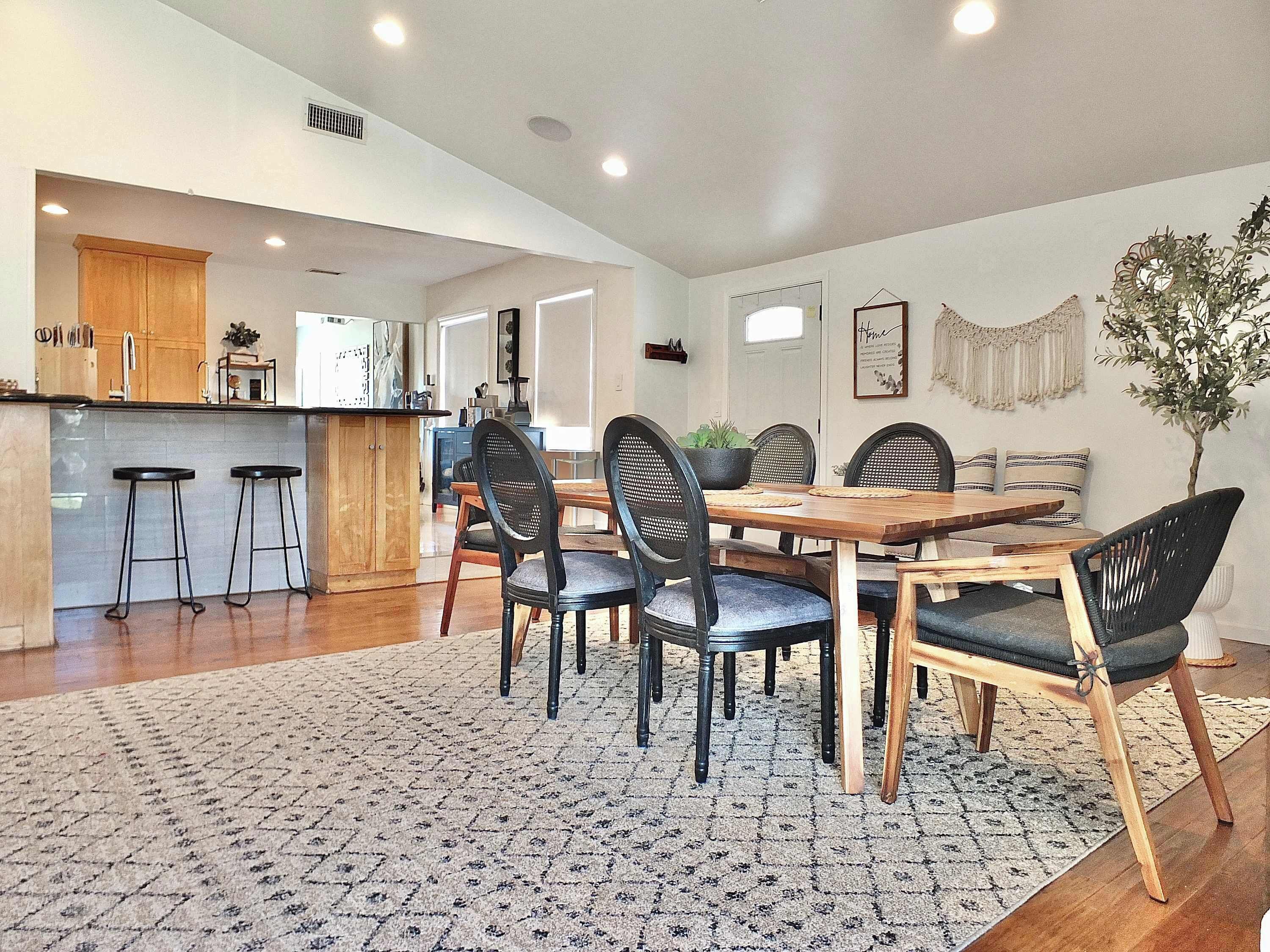 A spacious dining area with a wooden table and black chairs, a kitchen with wooden cabinets, and a patterned rug on the floor.