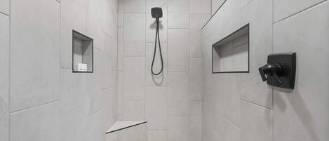 Una ducha moderna con cabezal de ducha de mano, panel de control en la pared y un espejo pequeño.