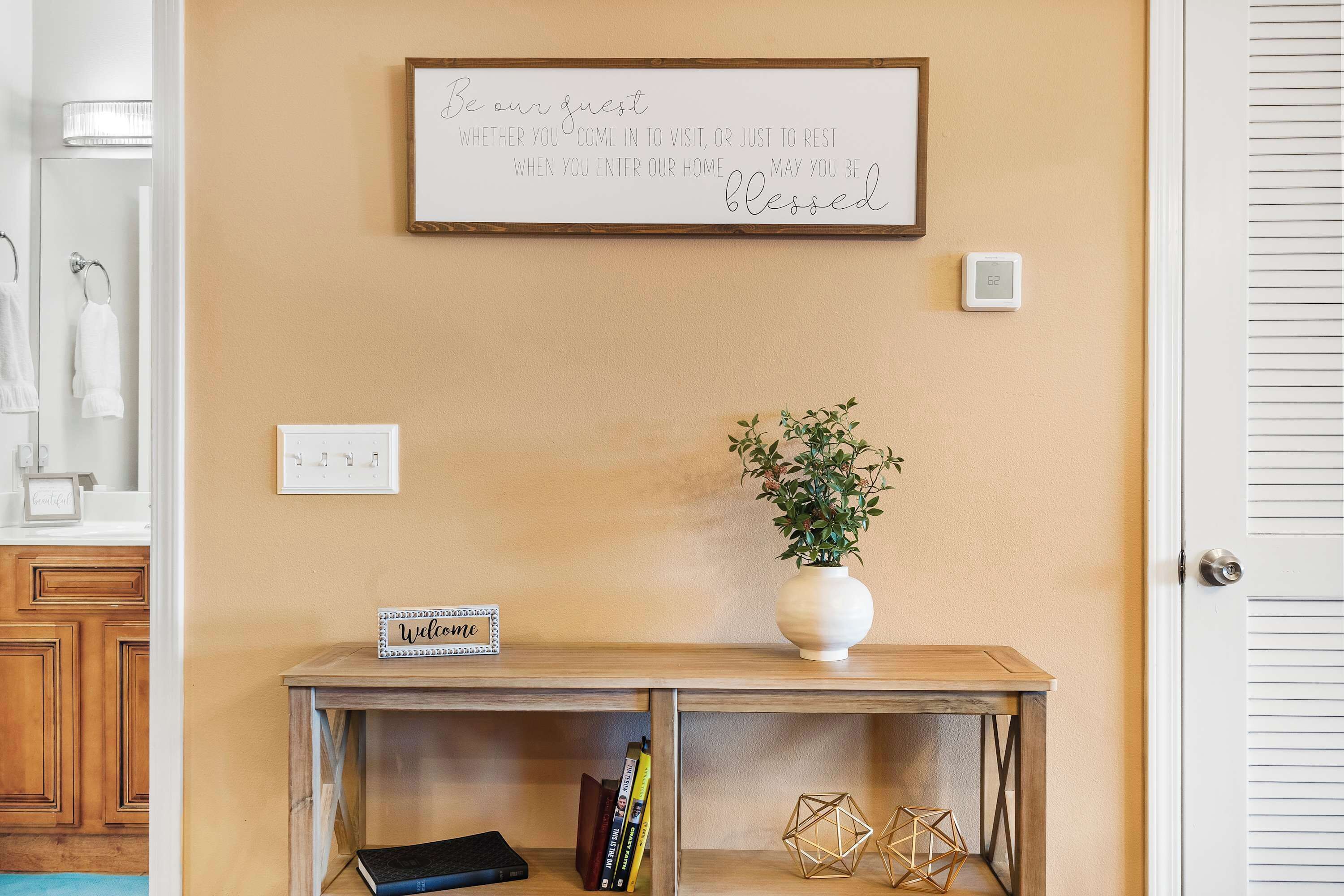 Un panneau fixé au mur avec un message d’accueil, une petite table sur laquelle est posé un panneau indiquant « Bienvenue », une plante en pot et un thermostat.