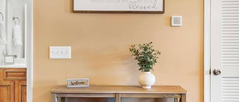 Un panneau fixé au mur avec un message d’accueil, une petite table sur laquelle est posé un panneau indiquant « Bienvenue », une plante en pot et un thermostat.