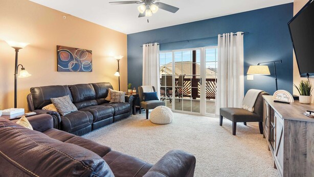 A living room with a black leather sofa, a ceiling fan, and a view of the outdoors through a sliding glass door.
