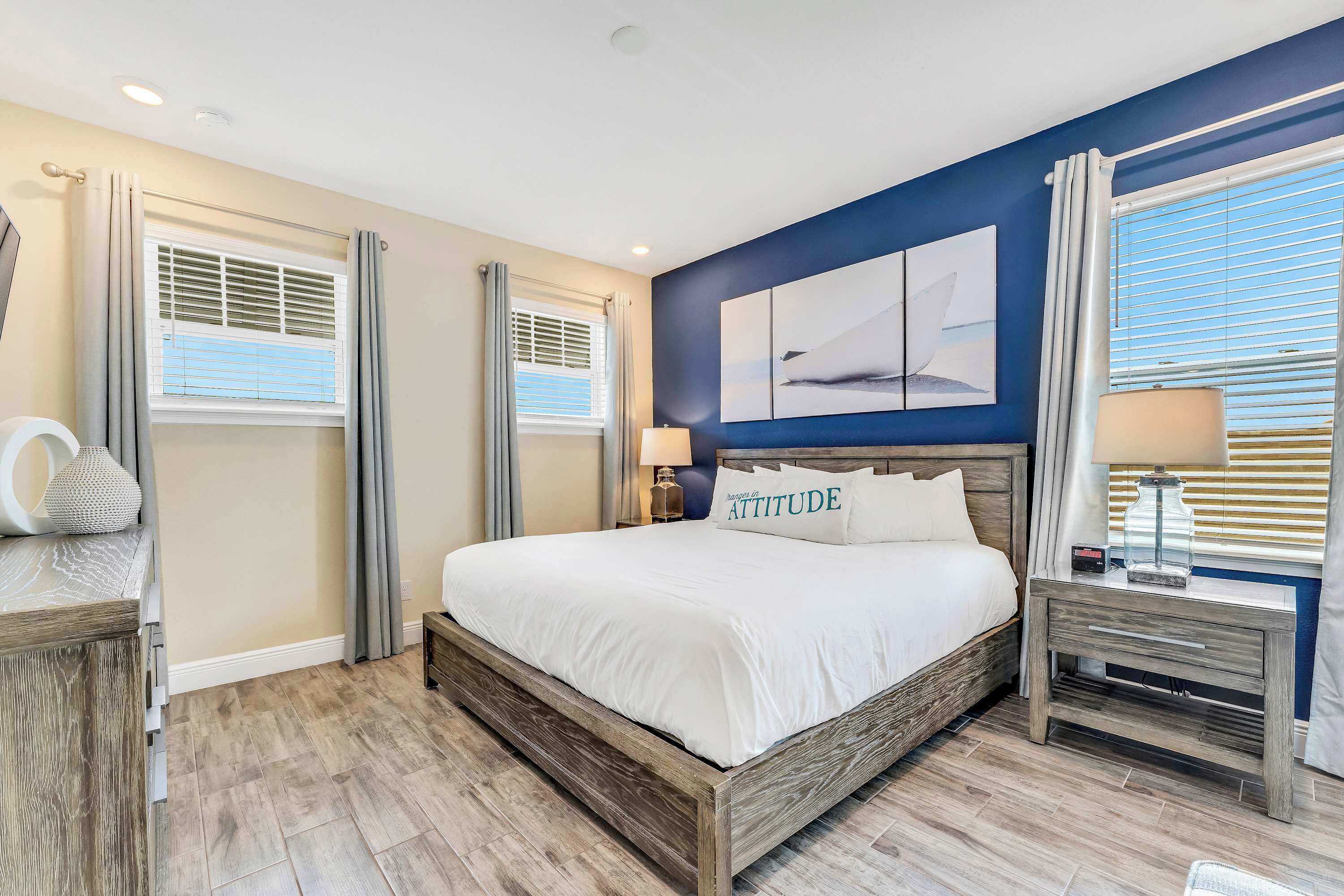A bedroom with a large bed, two bedside tables, a nightstand, a wall-mounted lamp, and a window with blinds.