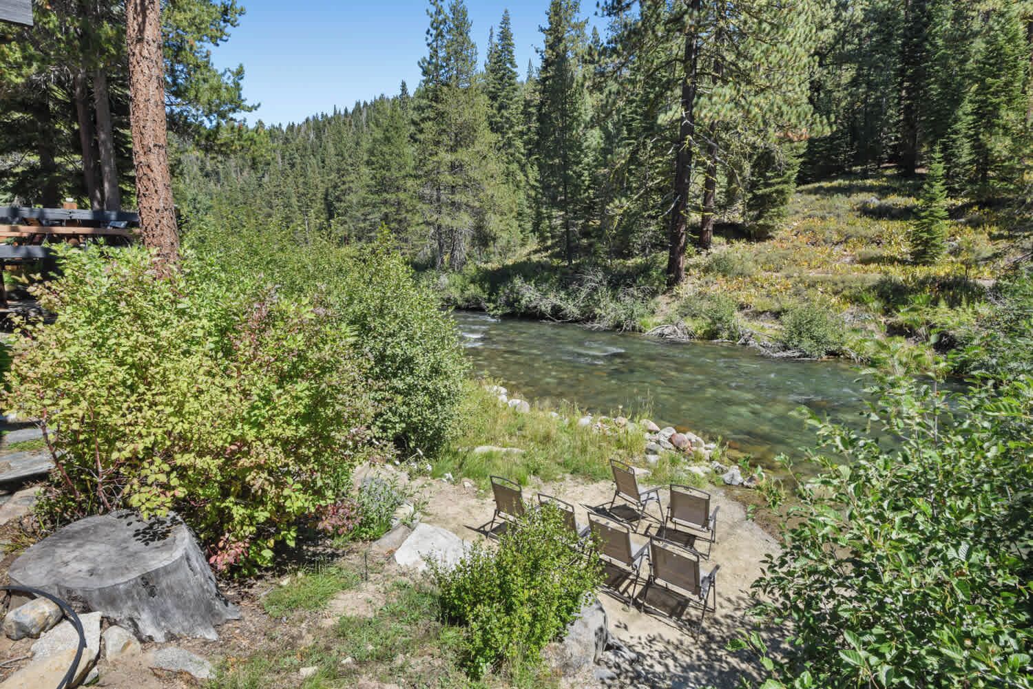 Un entorno junto al río con un banco y sillas para sentarse, rodeado de un bosque frondoso y un arroyo cristalino.