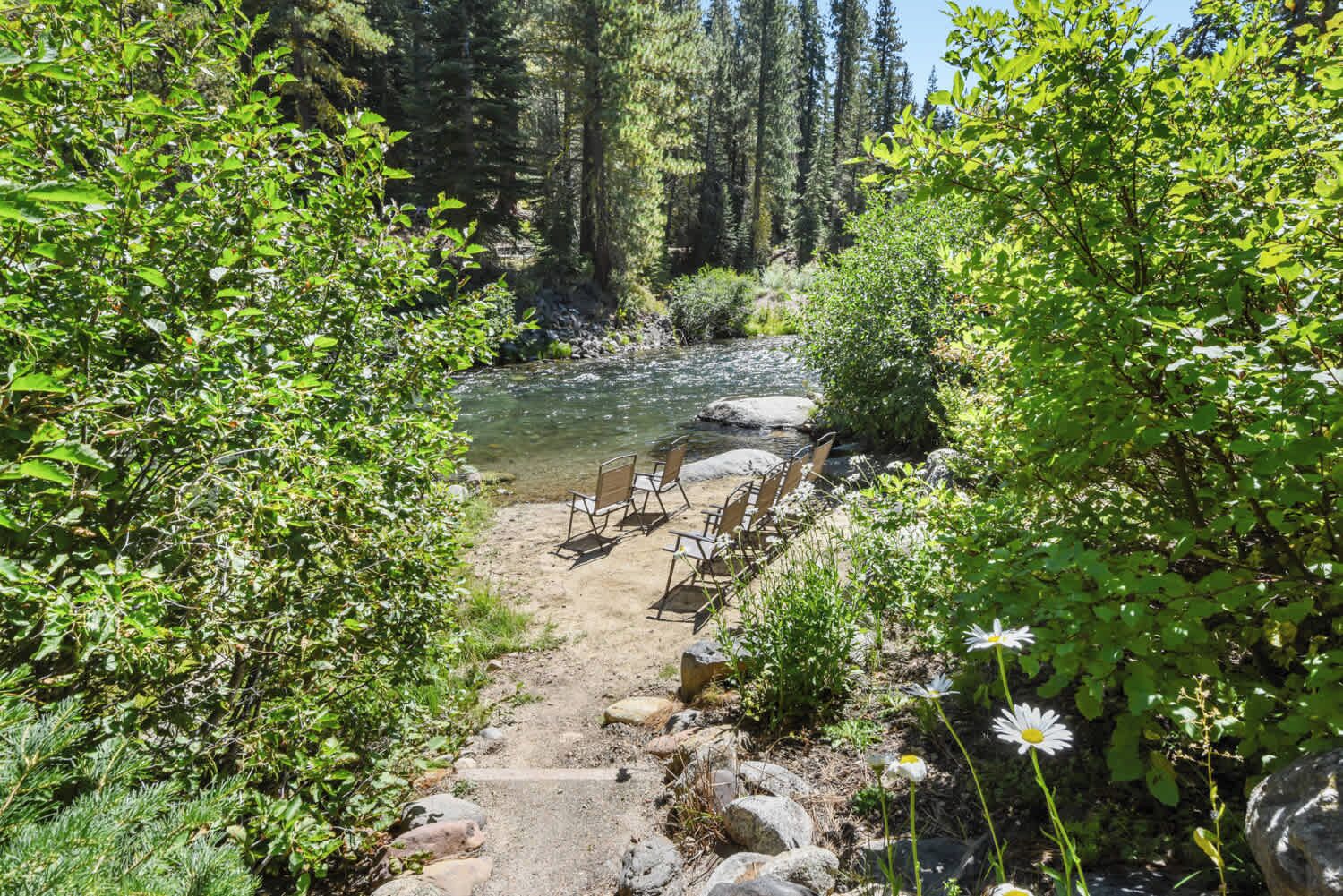 A natural setting with a clear stream, surrounded by dense greenery and rocks. There are wooden chairs placed along the streamside.