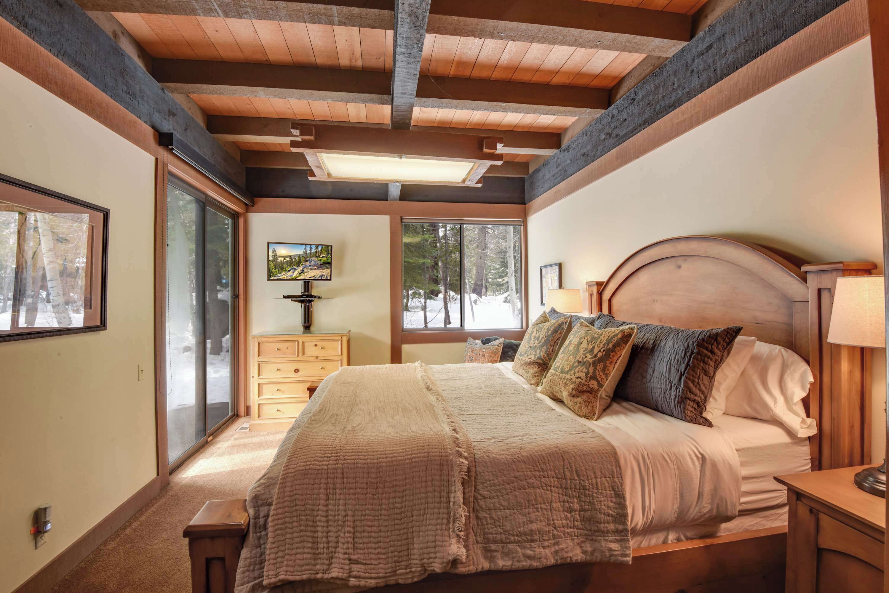 A bedroom with a wooden ceiling, a large bed with multiple pillows, a nightstand, a dresser, and a window with a view of trees.