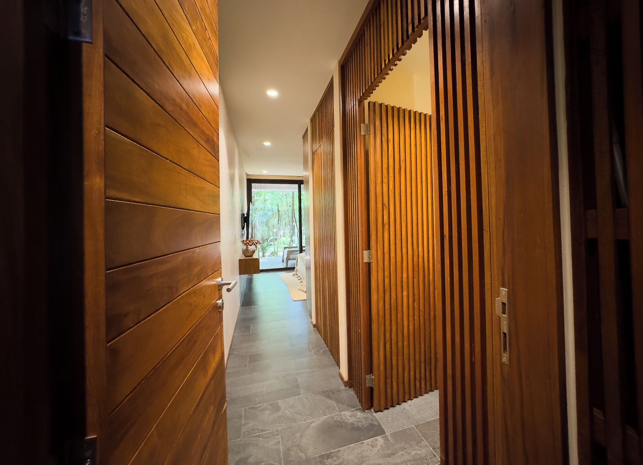 A hallway with wooden paneling and a stone floor leading to a room with a view of trees.
