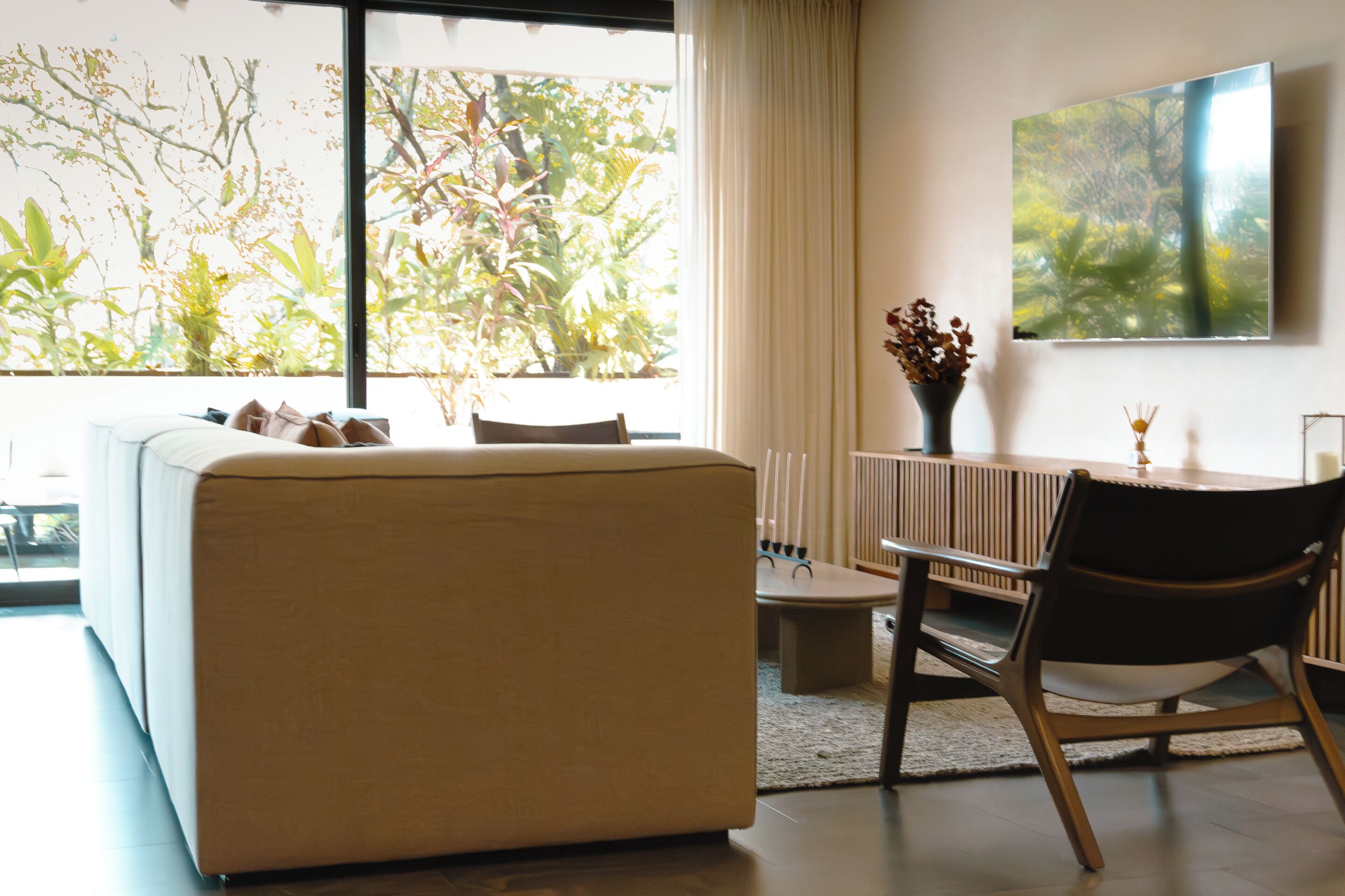 Uma sala de estar moderna com um sofá, uma mesa de jantar e uma cadeira. Há uma grande janela com vista para árvores e um quadro na parede.