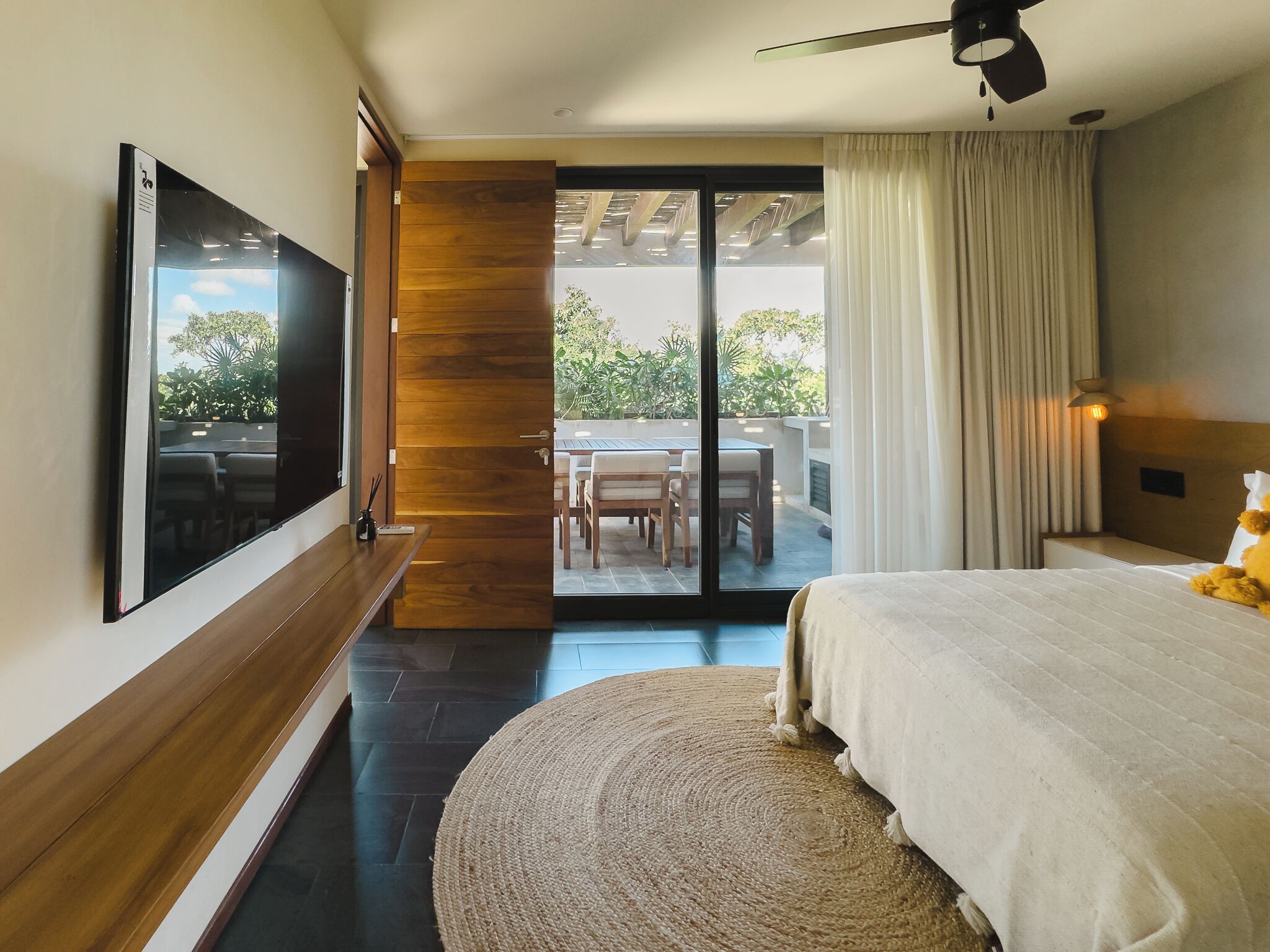 Ein Schlafzimmer mit Fernseher, Bett, einem runden Teppich, einer Holztür und Blick auf eine Terrasse.