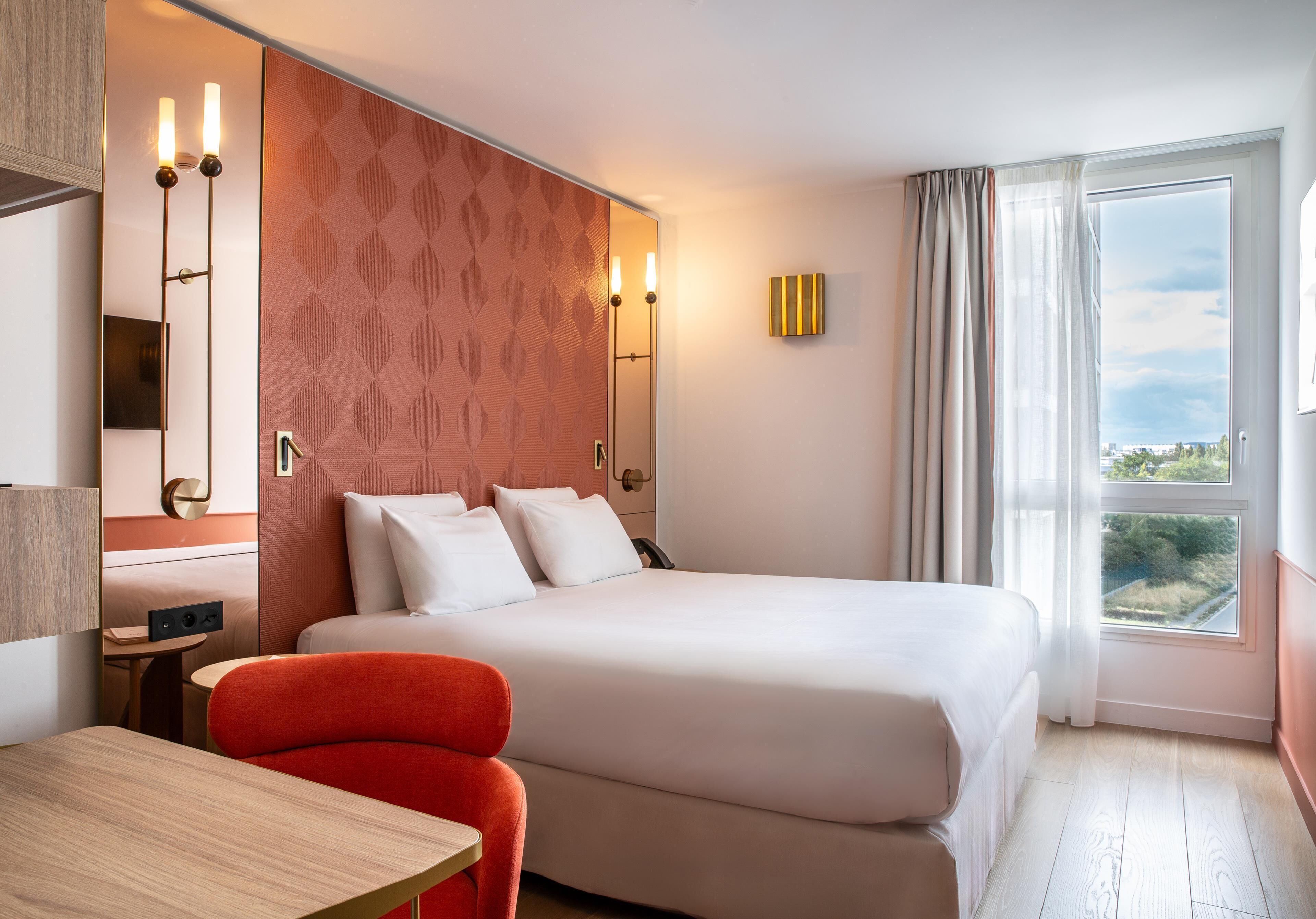 Une chambre d’hôtel comprenant un lit, un bureau, une chaise, une table de chevet, un miroir et une fenêtre avec des rideaux.