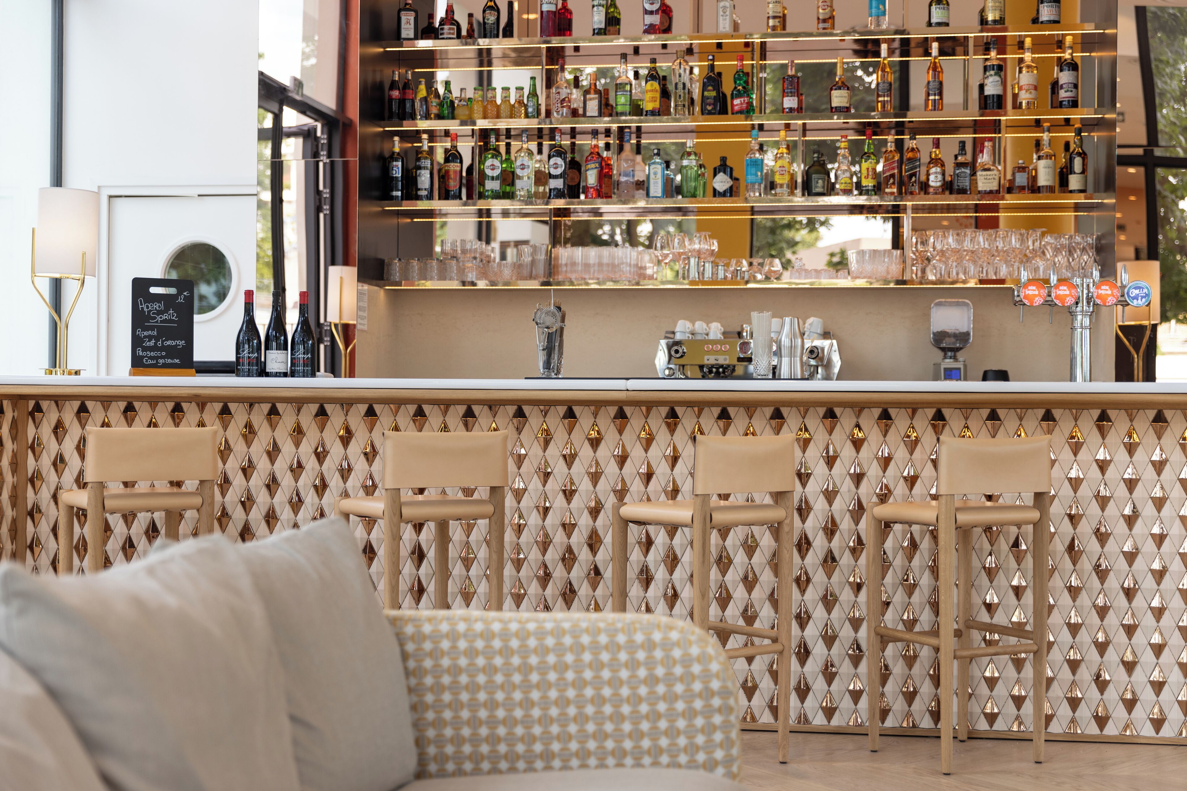 Un coin bar avec une variété de bouteilles d’alcool, des verres et une machine à café. On y voit trois tabourets et un canapé au premier plan.