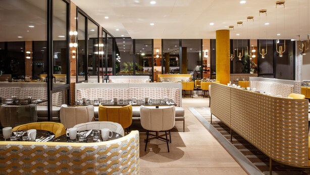 Een modern restaurant met glazen scheidingswanden, geel-witte inrichting en een centraal gelegen zitgedeelte.