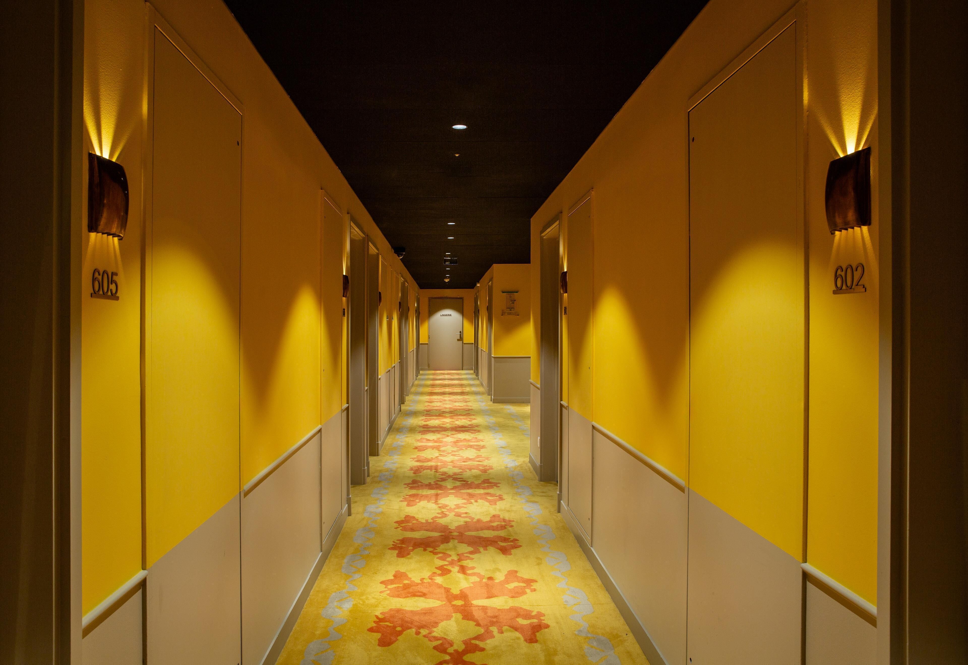 Un corridoio d'albergo con porte numerate 605 e 602, tappezzato con un motivo floreale, illuminato da luci a parete.