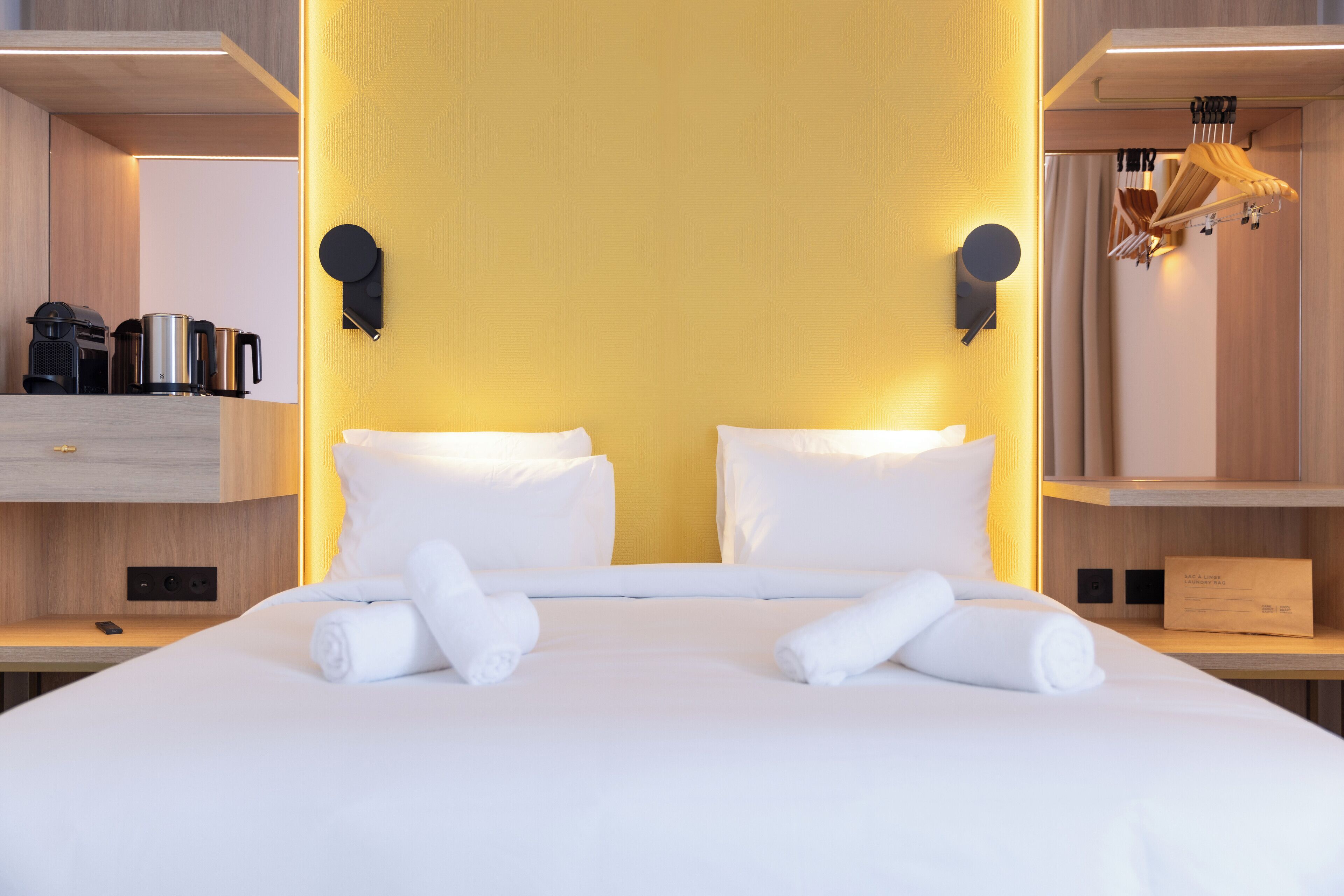 Cama bem arrumada com lençóis e toalhas brancos, cabeceira de madeira e parede amarela.