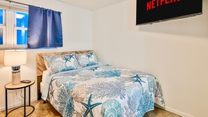 A bedroom with a bed, a nightstand, a lamp, a window with curtains, and a wall-mounted TV displaying "NETFLIX". - The Hideaway A Gulf Shores Dream Vacation! (Gulf Shores)