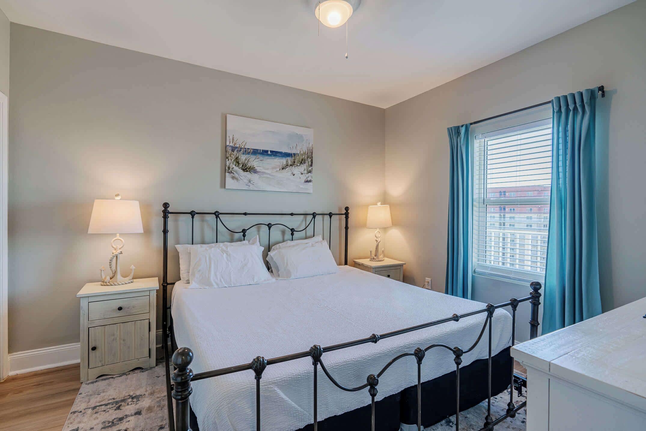 A bedroom with a metal bed frame, two bedside tables with lamps, a window with curtains, and a painting on the wall.