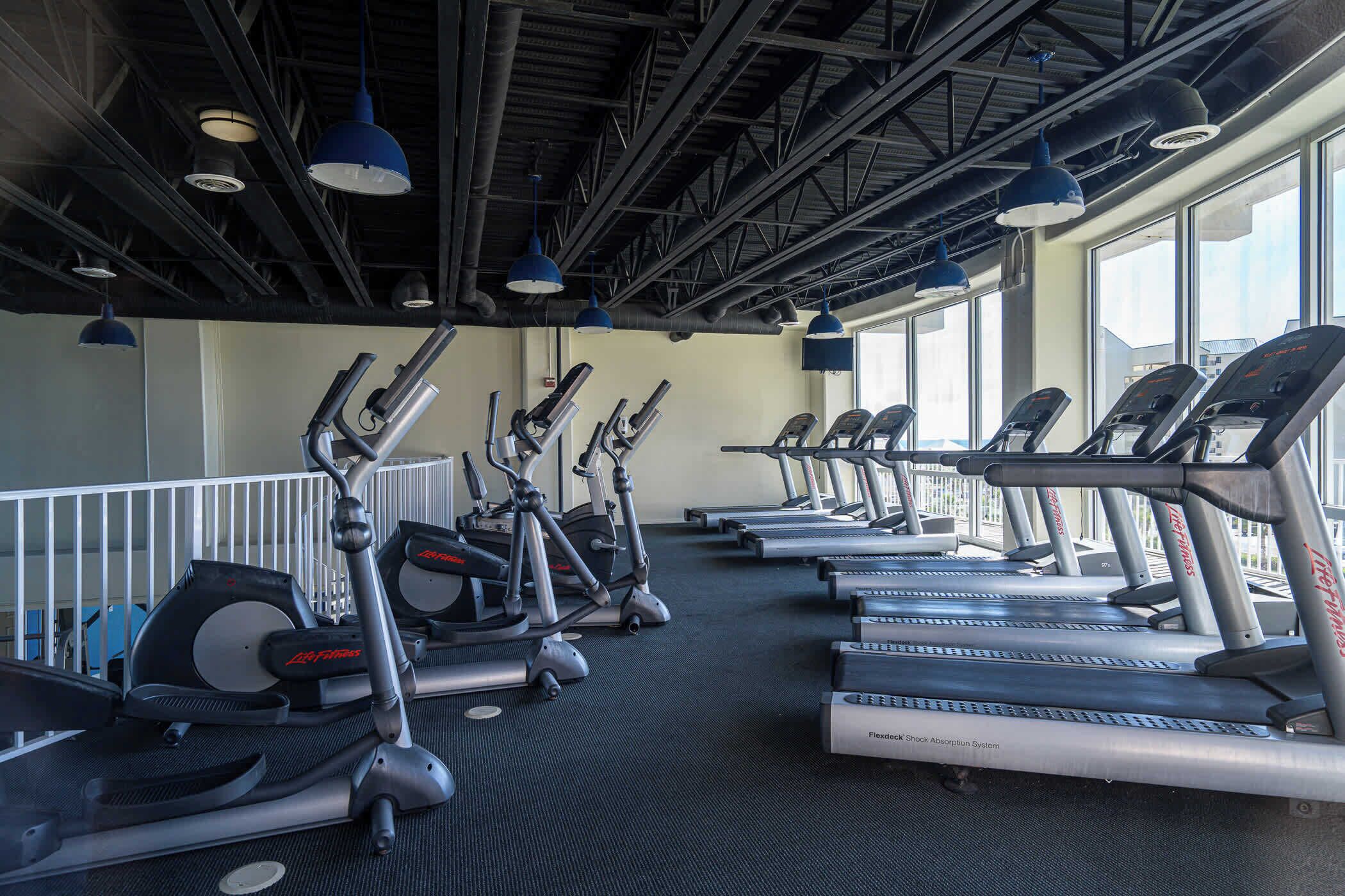 A gym with multiple treadmills, ellipticals, and a stationary bike, equipped with blue pendant lights and large windows.