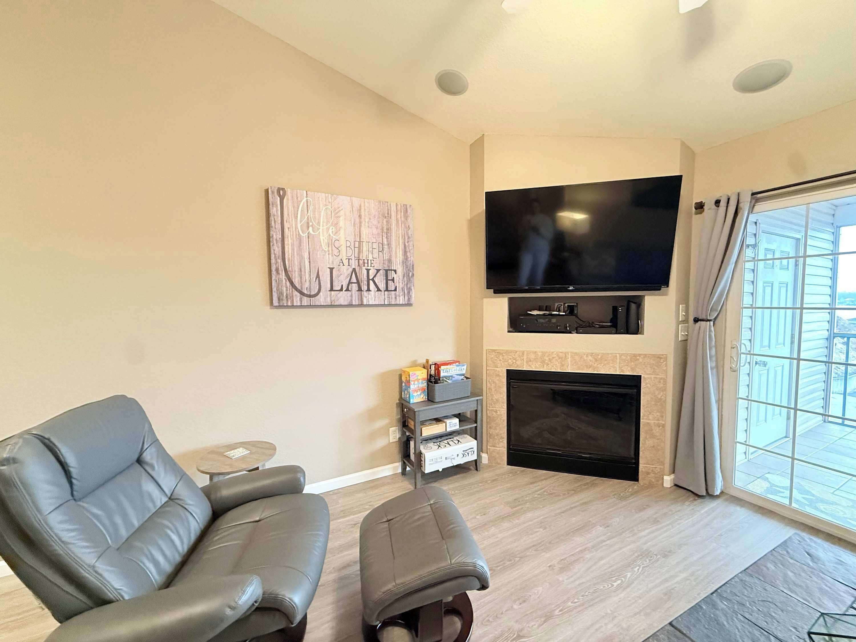 A living room with a grey leather sofa, a flat-screen TV mounted above a fireplace, and a window with curtains.