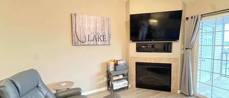 A living room with a grey leather sofa, a flat-screen TV mounted above a fireplace, and a window with curtains.