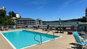 A swimming pool with lounge chairs and umbrellas, surrounded by a fence, with buildings and a lake in the background. - Highland Lake View Incredible Location! (Lake Ozark)