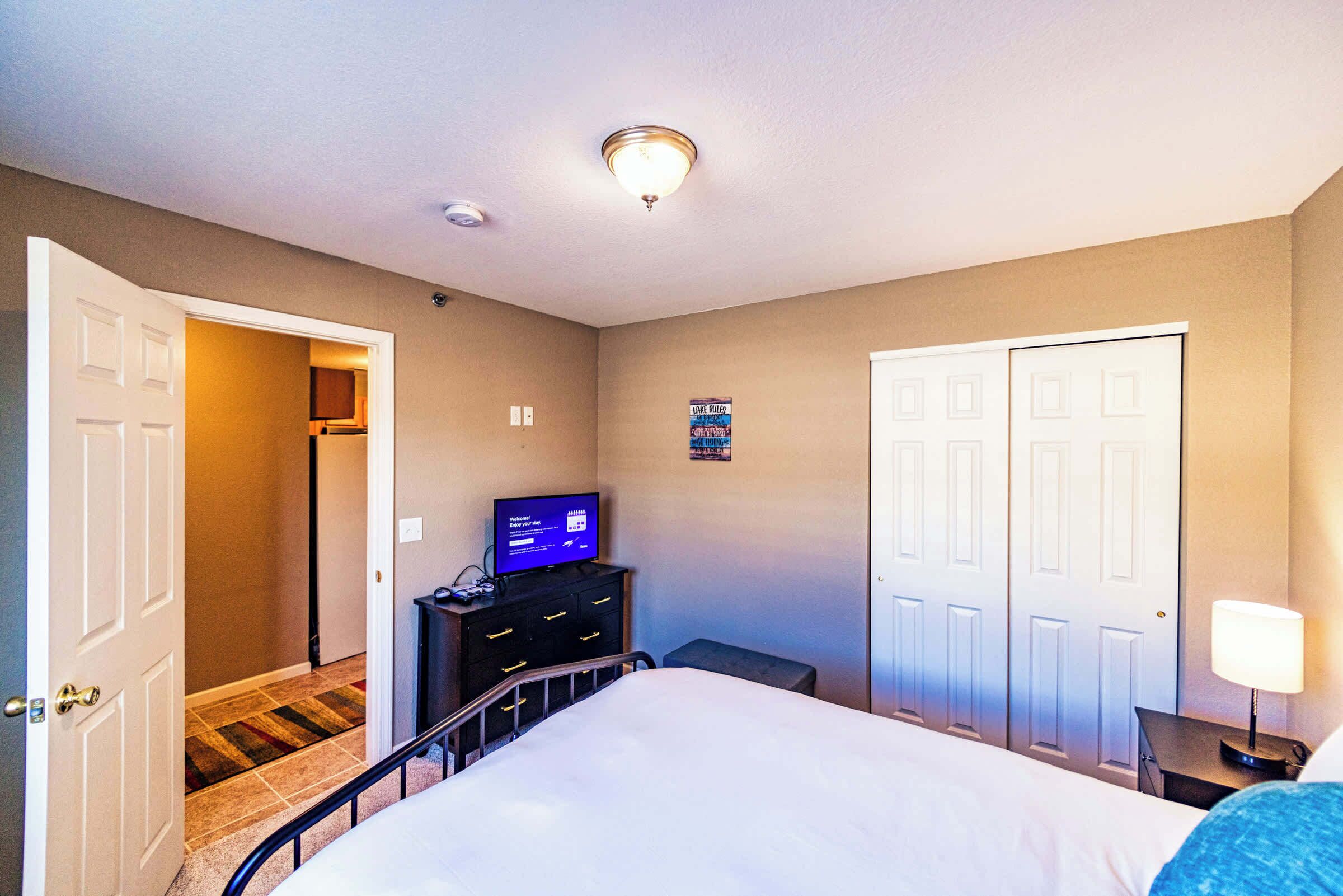 A hotel room with a bed, a television on a dresser, a door, and a wall-mounted light fixture.