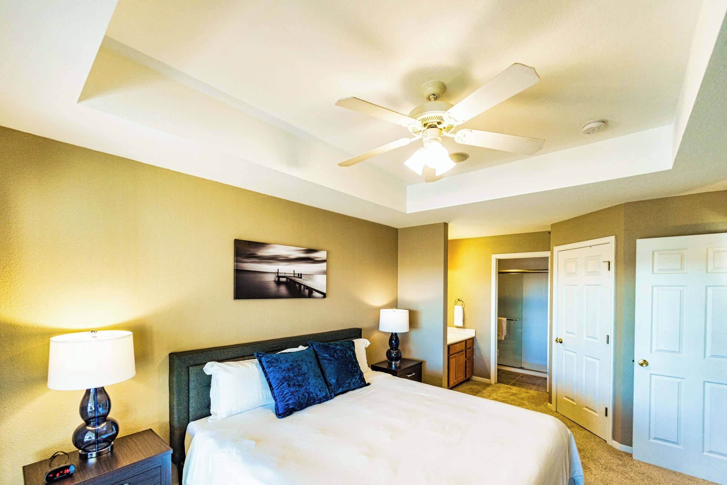 A hotel room with a bed, two bedside lamps, a ceiling fan, a painting on the wall, and a door leading to a bathroom.