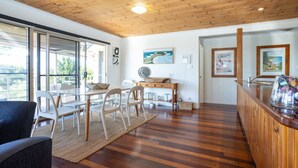 A modern interior with a wooden ceiling, a dining area with white chairs and a table, a kitchen counter with a basket, and artwork on the walls.