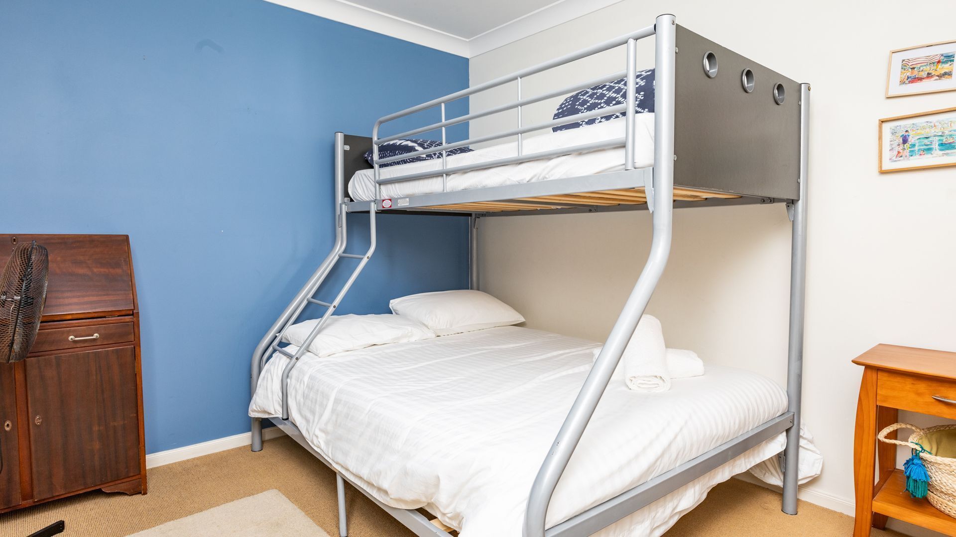 Un lit superposé dans une pièce aux murs bleus, avec une commode en bois et une petite table en bois sur laquelle se trouve un panier.