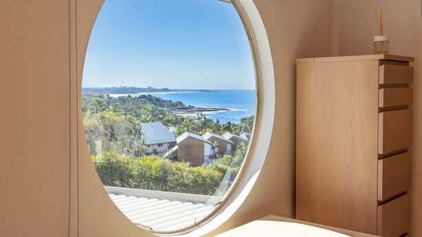 A room with a circular window offering a view of a coastal landscape, a wooden cabinet, and a small table.
