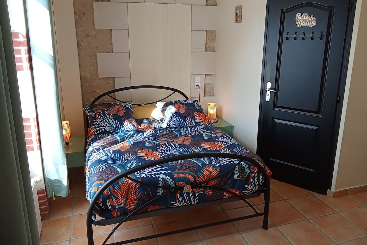 Un lit avec un cadre en métal, une couvre-lit à motifs, une étagère fixée au mur avec une lampe, et une porte avec une pancarte.