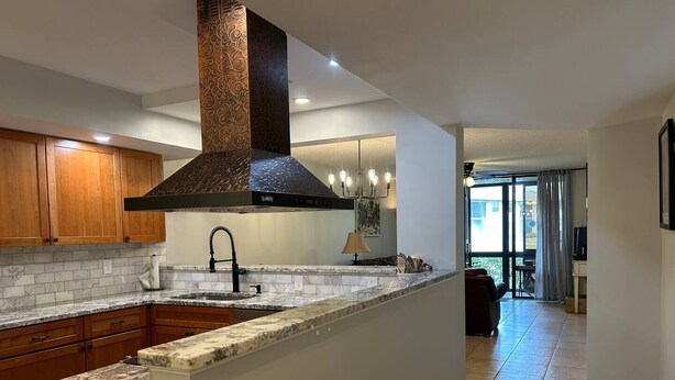 A modern kitchen with granite countertops, wooden cabinets, a stainless steel range hood, and a built-in island with a sink.