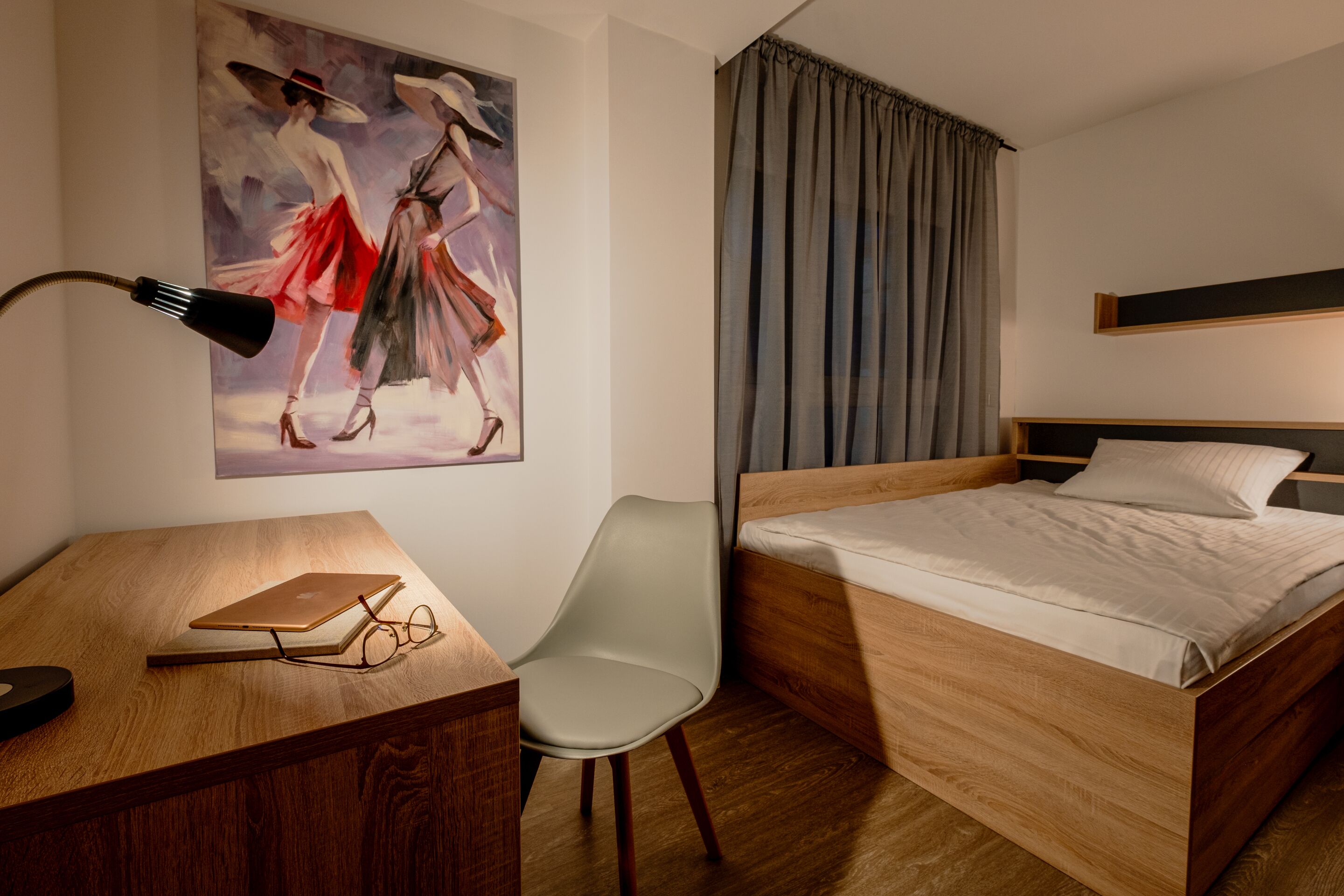 A hotel room with a wooden bed, a desk with a laptop, a chair, and a painting on the wall.