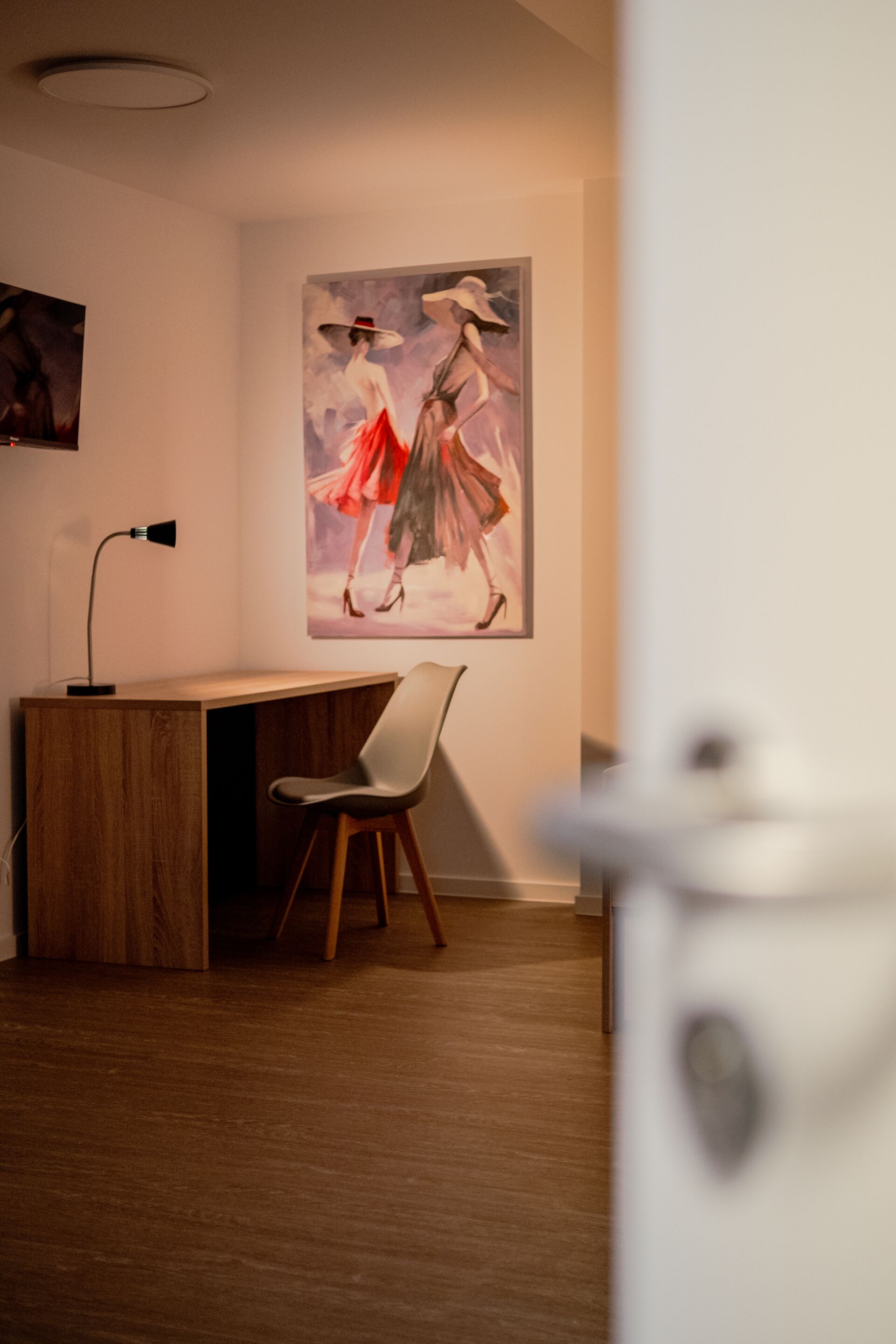 A room with a wooden desk, a white chair, and a painting on the wall.