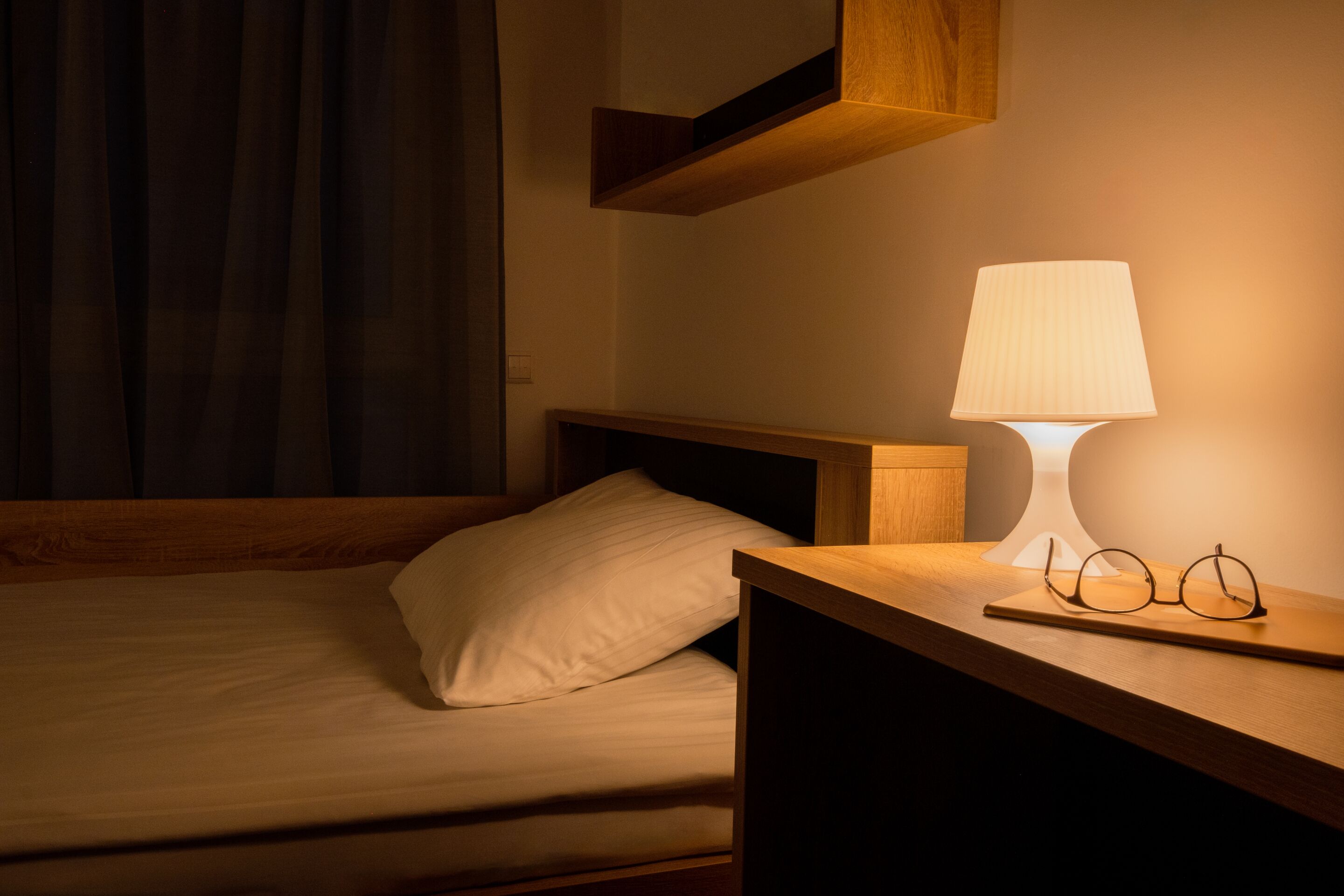 Ein ordentlich bezogenes Bett mit Kissen, ein Nachttisch mit Lampe und Gläsern sowie ein Holzregal darüber.