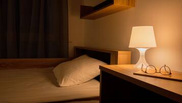 Ein ordentlich bezogenes Bett mit Kissen, ein Nachttisch mit Lampe und Gläsern sowie ein Holzregal darüber.