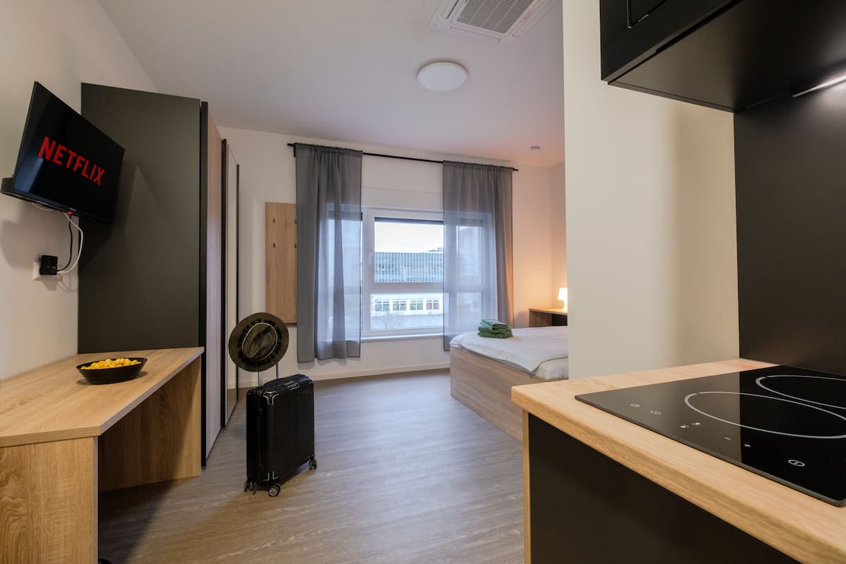 A modern hotel room with a bed, a desk, a TV, a suitcase, and a kitchen area with a stove.