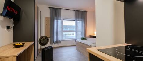 A modern hotel room with a bed, a desk, a TV, a suitcase, and a kitchen area with a stove.