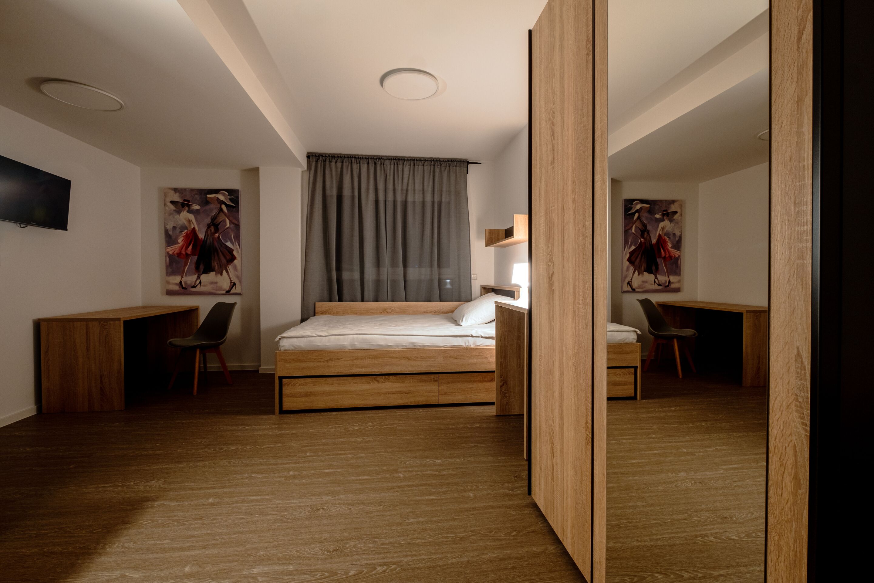 Un dormitorio moderno con una cama de madera, un escritorio y una silla. Hay dos cuadros en las paredes, una lámpara en el techo y una ventana con cortinas.