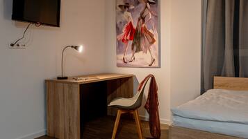 Habitación de hotel con un escritorio de madera, una silla, una cama, un televisor montado en la pared y un cuadro con la imagen de dos personas.