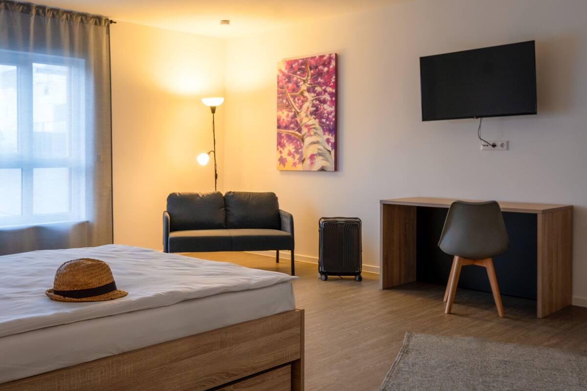 A hotel room with a bed, a desk, a chair, a lamp, a TV, and a painting on the wall.